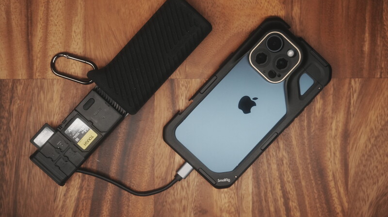 iPhone with rugged case connected to portable power bank and card reader on wooden surface.