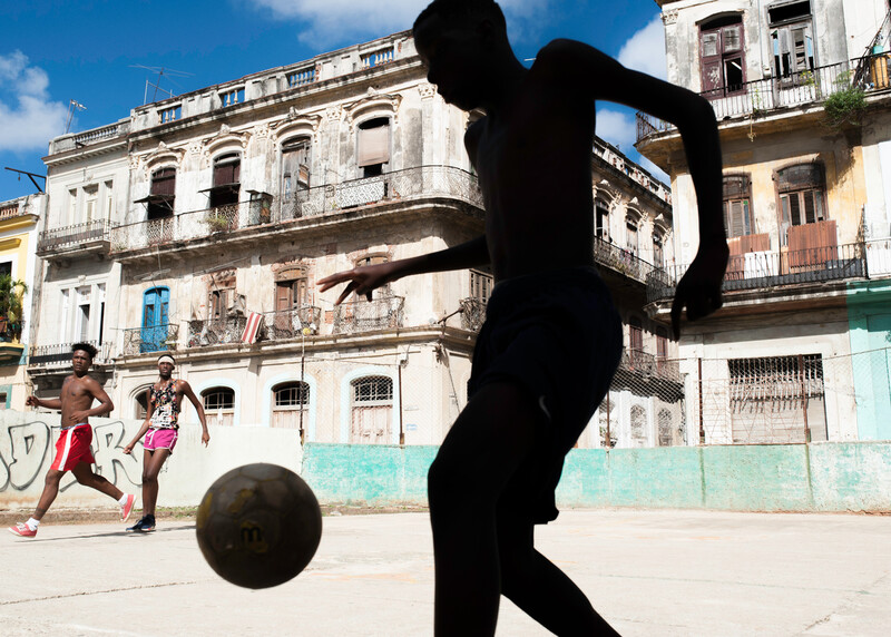 Silhouetted soccer player performing an athletic kick with ball, colorful colonial buildings in background.