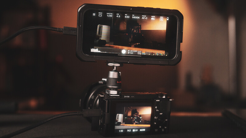 A smartphone mounted on a tripod displaying live video feed above a professional cinema camera, both showing the same golden-hour interior scene.