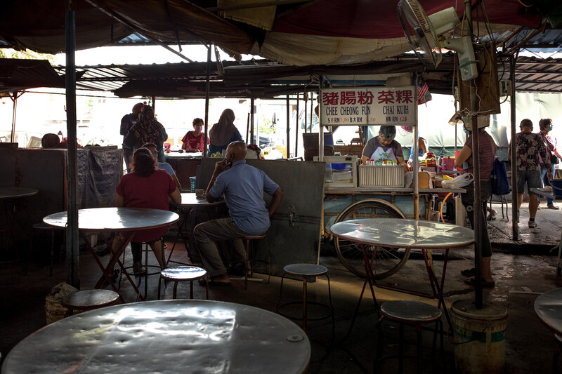 Interior of a covered outdoor market or food hall with patrons seated at round tables under natural light.