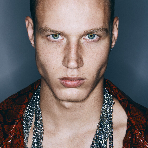 Portrait of a man with pale eyes against a dark background, wearing a red patterned jacket.
