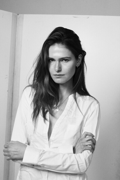 Woman in white button-up shirt against white wall, posed with contemplative expression.