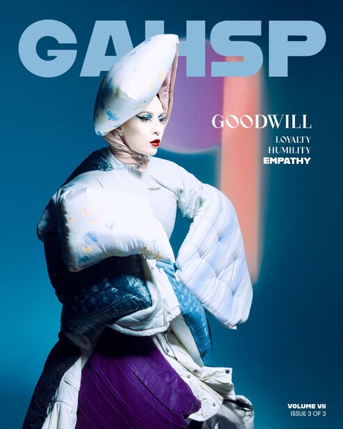 Person wearing sculptural white headpiece and draped white garment against blue and purple background.