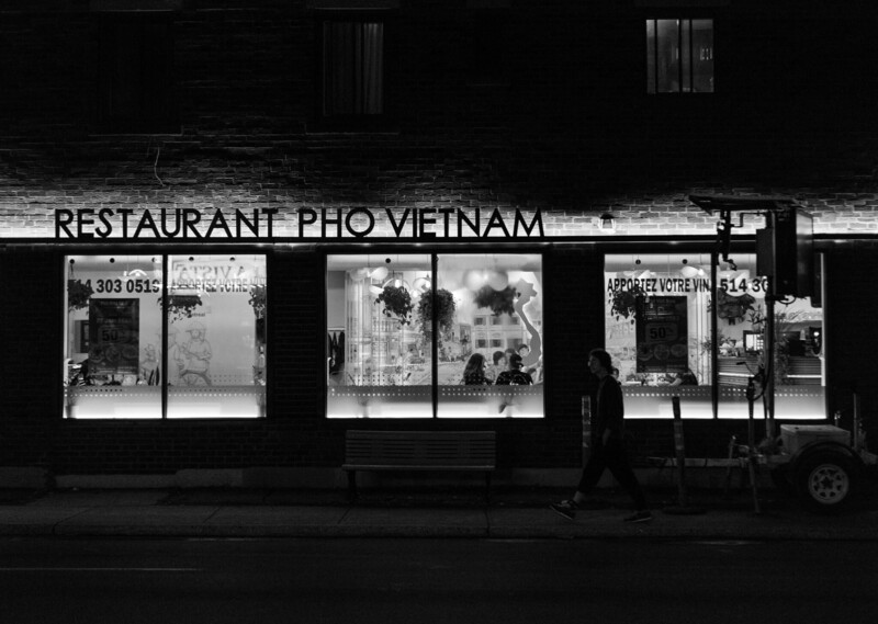 Street-level storefront of a Vietnamese pho restaurant at night, captured in high-contrast black and white.
