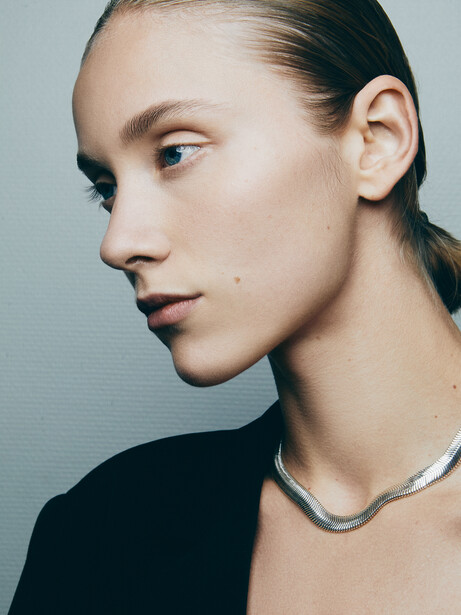 Woman in profile wearing a silver chain necklace against a neutral blue-gray background.