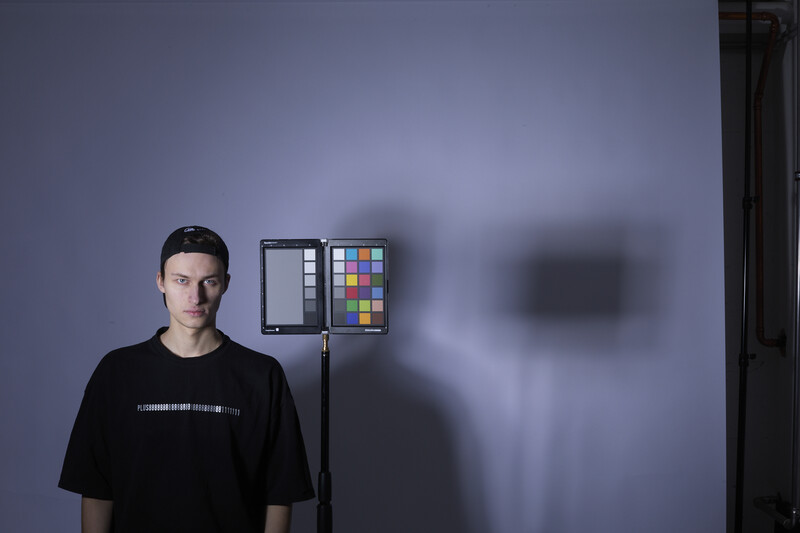 Portrait of a young man standing against a blue wall next to a color checker chart mounted on a light stand.