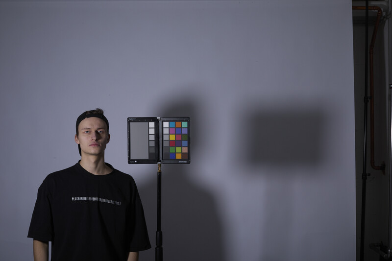 Man in black t-shirt standing beside studio lighting setup with color reference chart on gray backdrop.