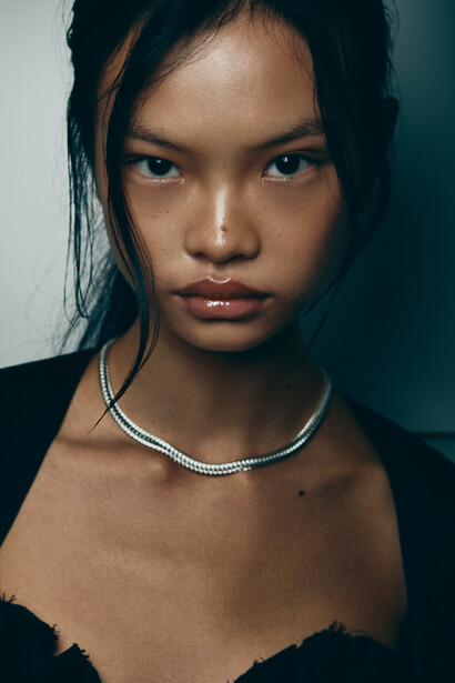 Portrait of a young woman with dark hair and a silver chain necklace against a teal background.