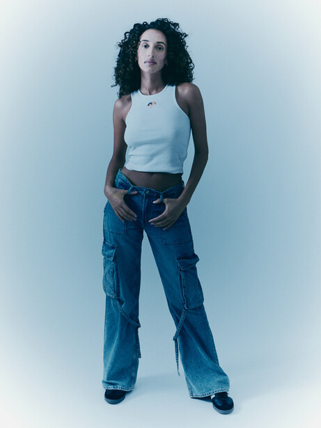 Woman in light blue tank top and blue cargo jeans standing against neutral backdrop.