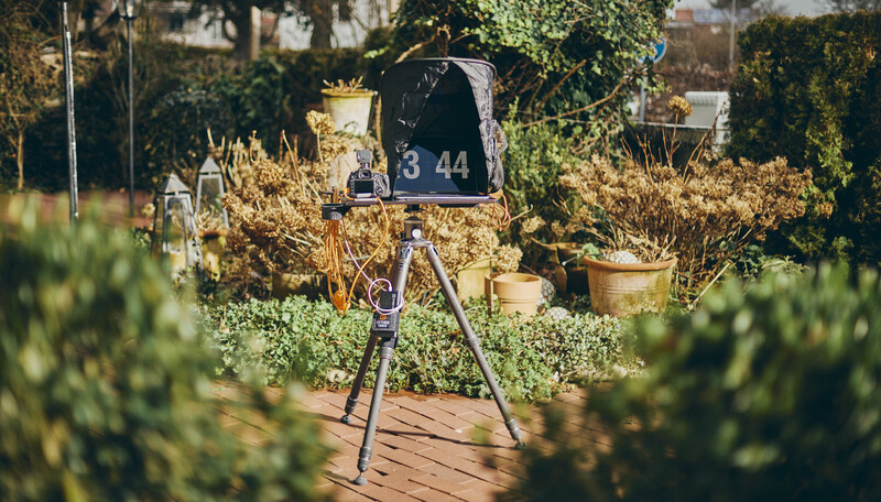 Film camera mounted on tripod in a garden surrounded by dried plants and potted herbs.