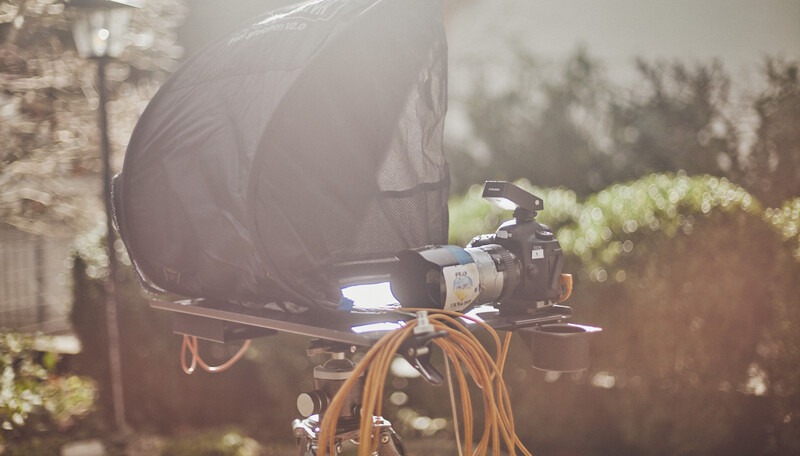 Professional video camera mounted on tripod with large black soft box reflector and orange cables in outdoor garden setting.