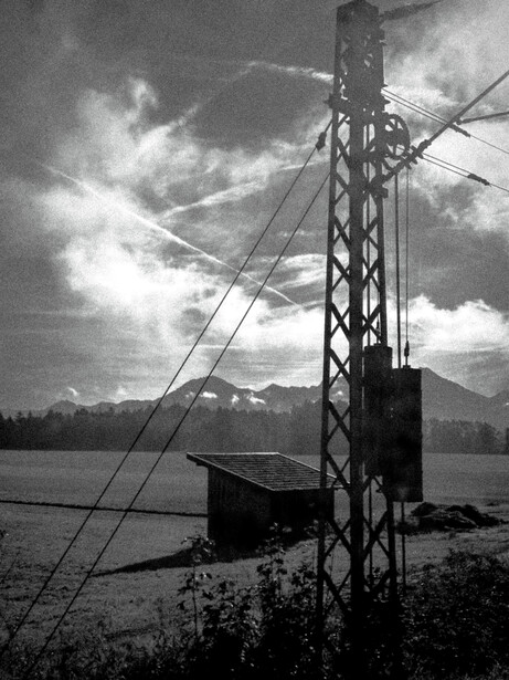 Tall steel transmission tower silhouetted against dramatic cloudy sky with mountain and water in background.