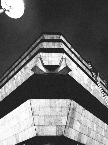 Upward view of brutalist concrete building with stacked chevron-shaped levels against dark sky and moon.