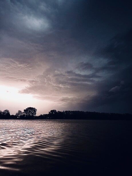Dramatic storm clouds gathering over still water at dusk, silhouetted trees lining the distant shoreline.
