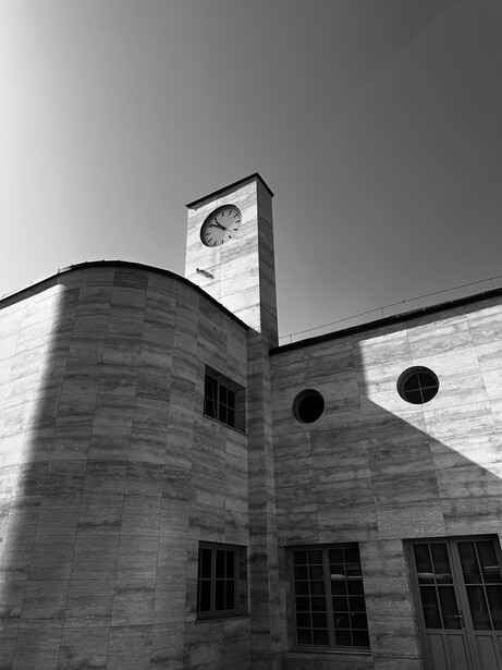 Modernist concrete clock tower rising above cylindrical building with circular windows in black and white.