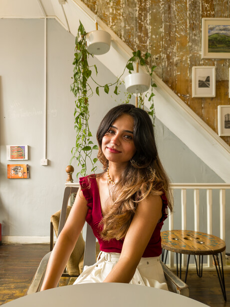 Woman with long dark hair seated at a table, wearing a red sleeveless top and white skirt, in a bright studio or home interior.