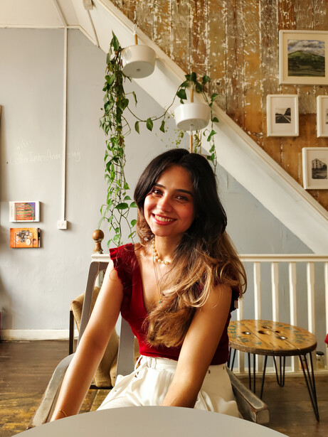 Woman in red sleeveless top seated indoors, smiling at camera with staircase and decorative plants in background.