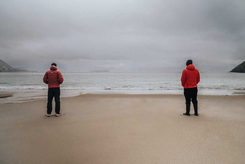 Two people in red jackets standing separately on a beach facing the ocean under overcast skies.