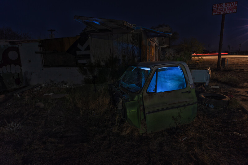 Abandoned green truck with glowing blue interior parked in a junkyard at dusk.