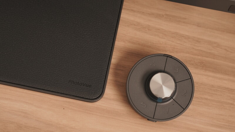 Black wireless control device with metallic center knob on wooden desk surface next to dark mousepad.