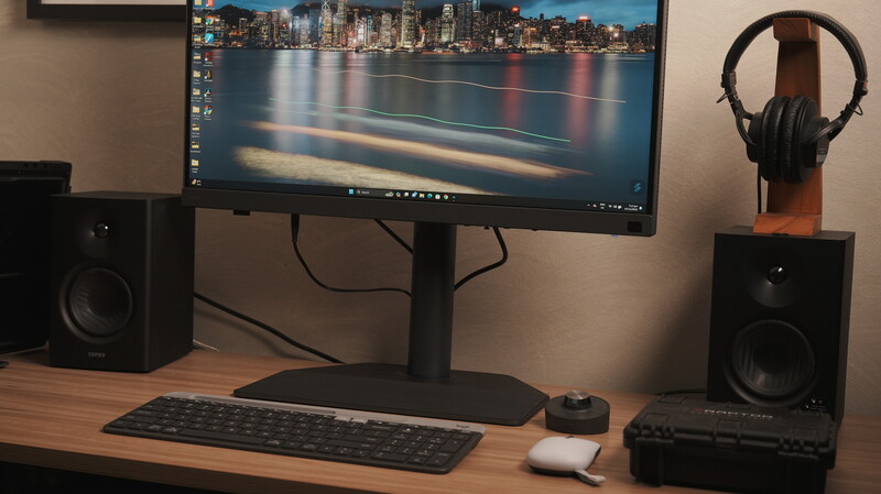 Photography workspace with monitor displaying cityscape long exposure, keyboard, mouse, studio monitors, and headphones on wooden desk.
