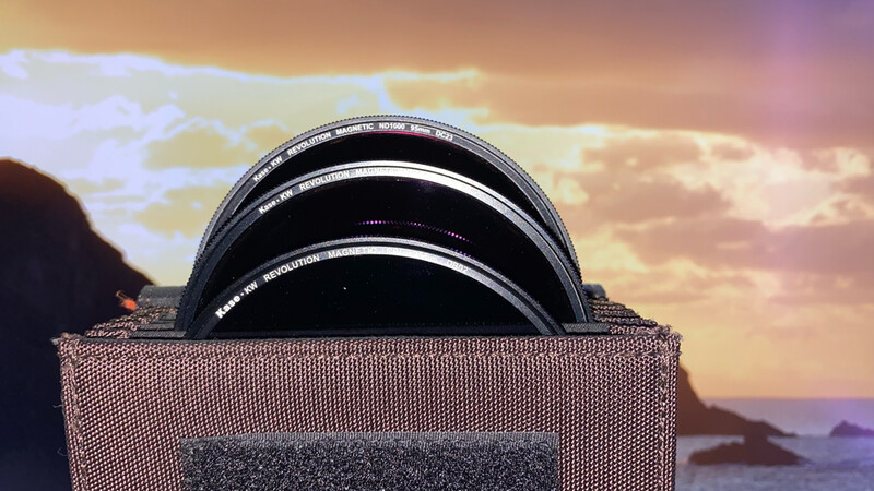 Stack of camera lens filters in a mesh pouch against a dramatic sunset sky over water.