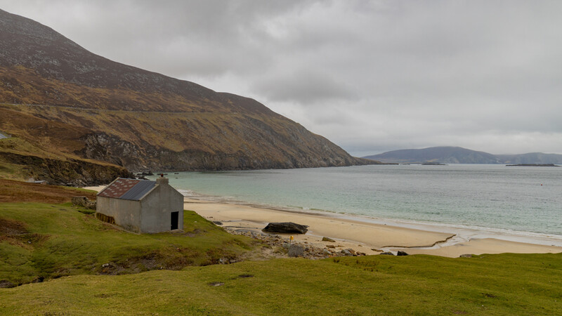 Abandoned white cottage on grassy slope overlooking a sandy beach and turquoise bay beneath clouded sky.