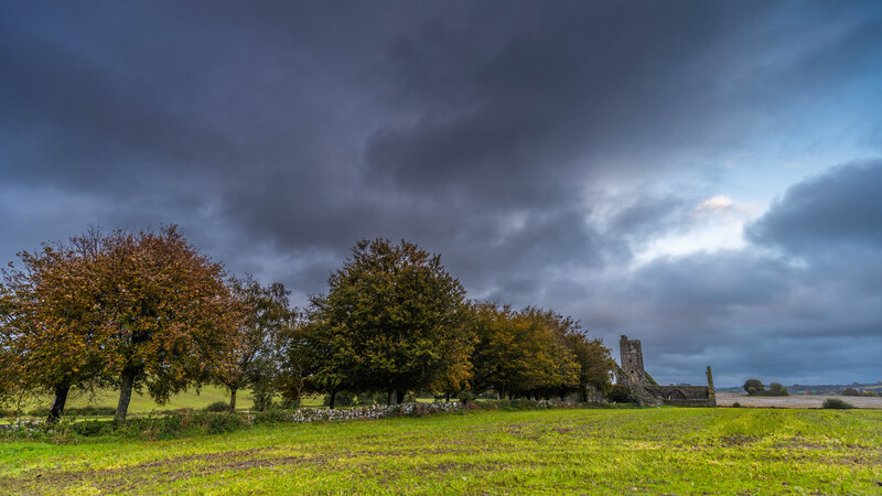 Pastoral landscape with autumn trees and distant church ruins beneath dramatic storm clouds.