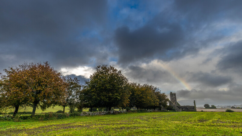 Rural landscape with mature trees and stone ruins under dramatic cloudy sky.