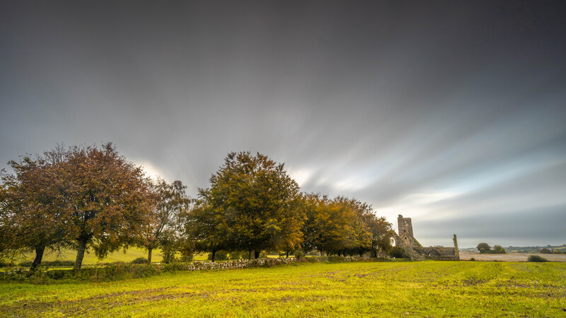Rural landscape with autumn trees and distant stone tower under dramatic cloudy sky.