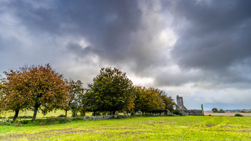 Rural landscape with scattered trees and distant church ruins under dramatic cloudy sky.