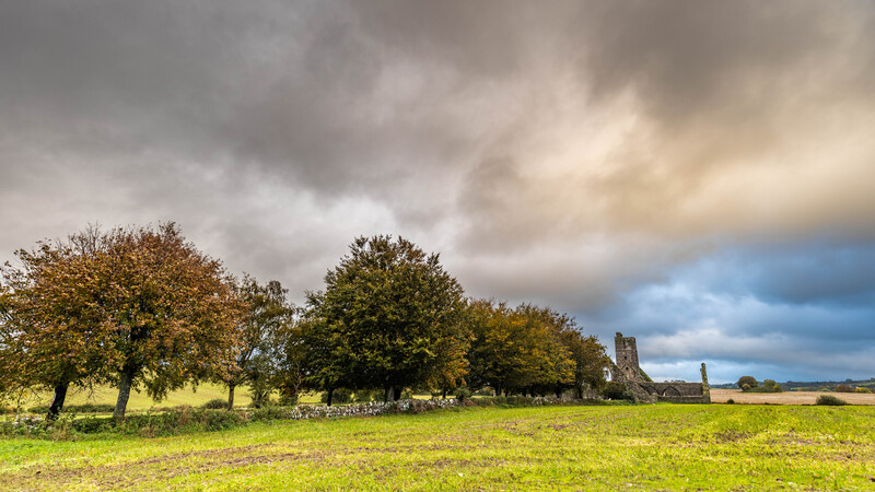 Pastoral landscape with mature trees and ruined stone structure under dramatic cloudy sky.