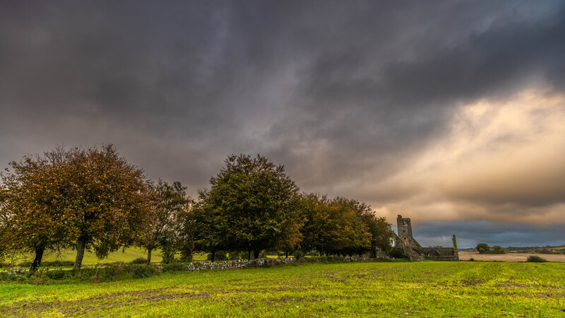 Dramatic stormy sky over rural grassland with scattered trees and distant stone ruins.