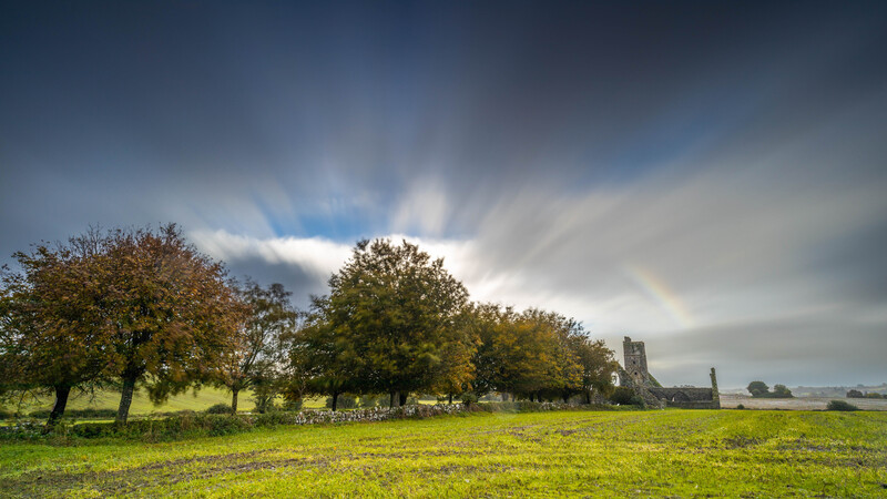 Rural landscape with scattered trees and distant ruins beneath dramatic cloudy sky.