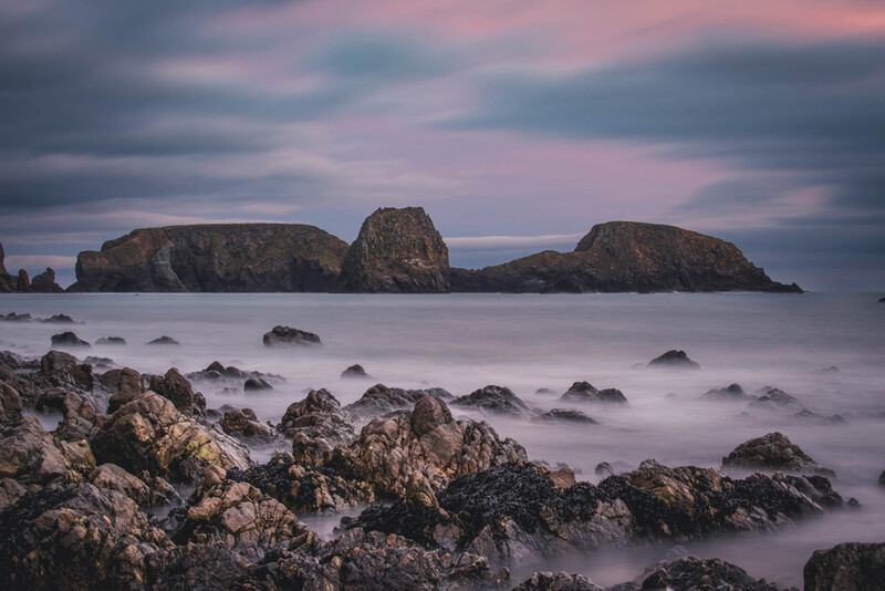 Rocky coastline with misty seawater and rugged islands under a moody twilight sky.