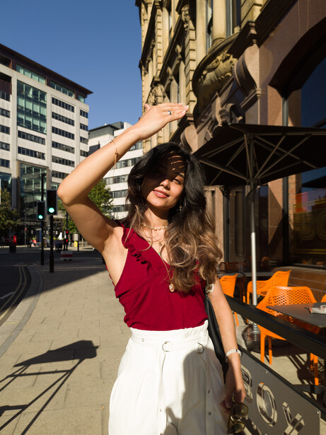 Woman in red halter top and white pants posing under a storefront awning in bright sunlight.
