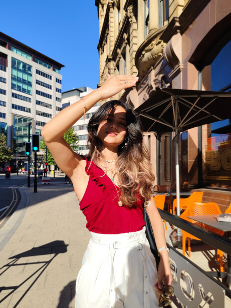 Woman in red top and white pants shading her eyes while looking upward on a sunny urban street.
