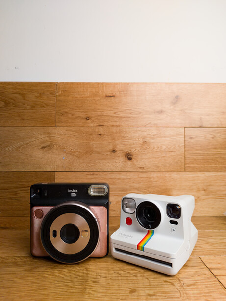 Two instant film cameras on wooden flooring: a brown and black vintage model and a white model with rainbow stripe detail.