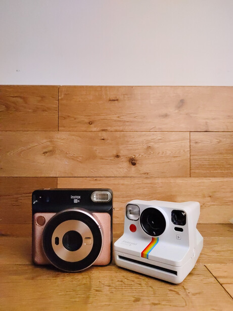 Fujifilm Instax SQ6 and Polaroid Now instant cameras side by side on wooden floor.