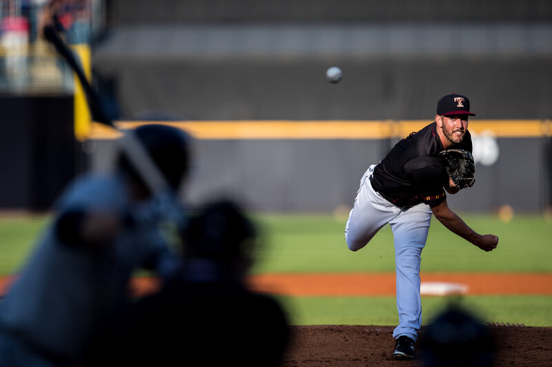 Baseball pitcher mid-delivery with sharp focus on the athlete and blurred batter and catcher in foreground.