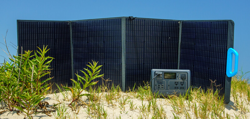 Portable solar panel array with digital power controller on sandy beach with coastal vegetation.