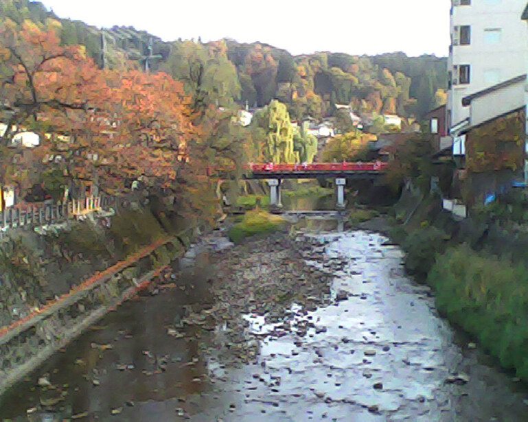 River flowing through an urban valley surrounded by autumn foliage and buildings, with a red pedestrian bridge spanning the water.