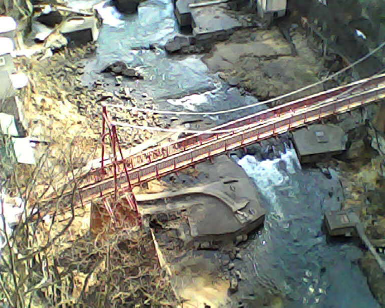 Aerial view of a red metal bridge spanning a muddy waterway with surrounding terrain and vegetation.