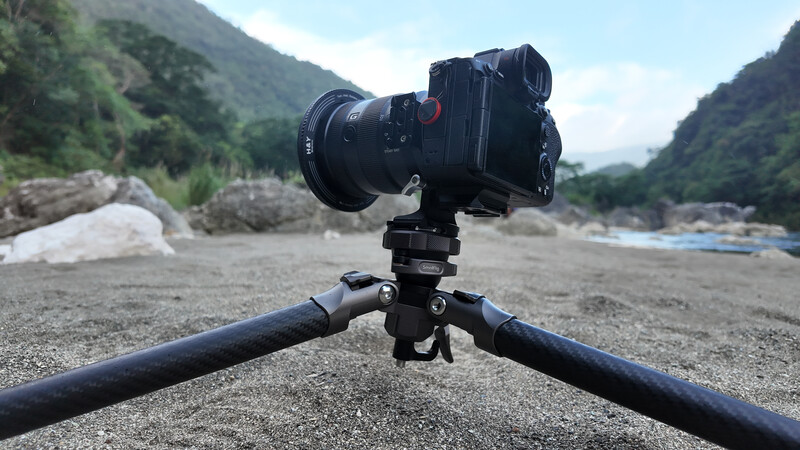 Professional mirrorless camera mounted on tripod legs at a rocky riverbed with forested mountains in background.