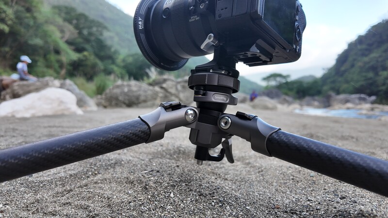 Video camera mounted on a tripod with spread legs positioned on sandy ground, with forested hills and a river valley in the background.