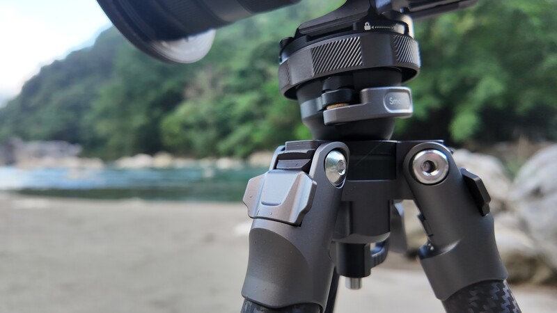 Professional tripod head with ball mount positioned on a beach with forested cliffs and water in the background.
