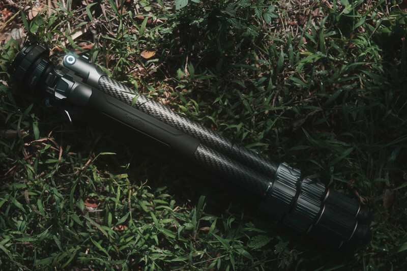 Black telephoto lens with carbon fiber texture lying diagonally on green grass and foliage.
