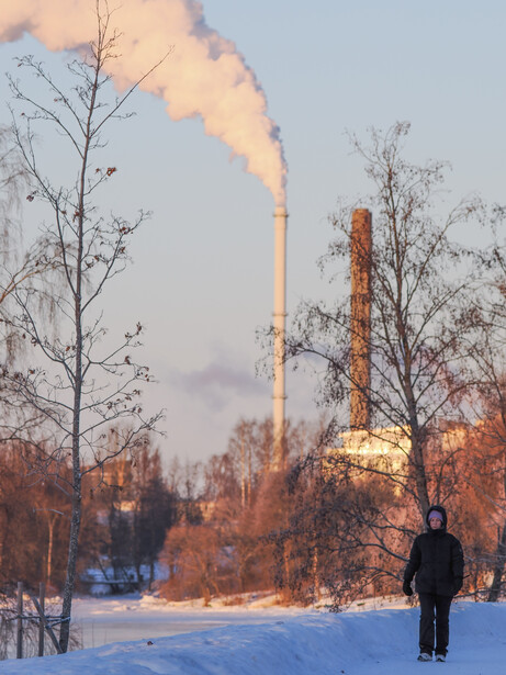 Person standing in snowy landscape facing industrial smokestacks emitting plumes of smoke at dusk.