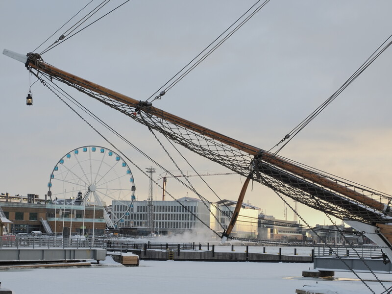 Historic sailing ship's wooden boom and rigging silhouetted against a waterfront cityscape with a ferris wheel.