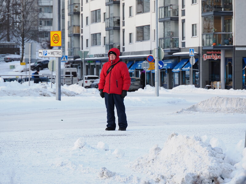 Person in red jacket standing in snowy urban plaza with apartment buildings and storefronts in background.